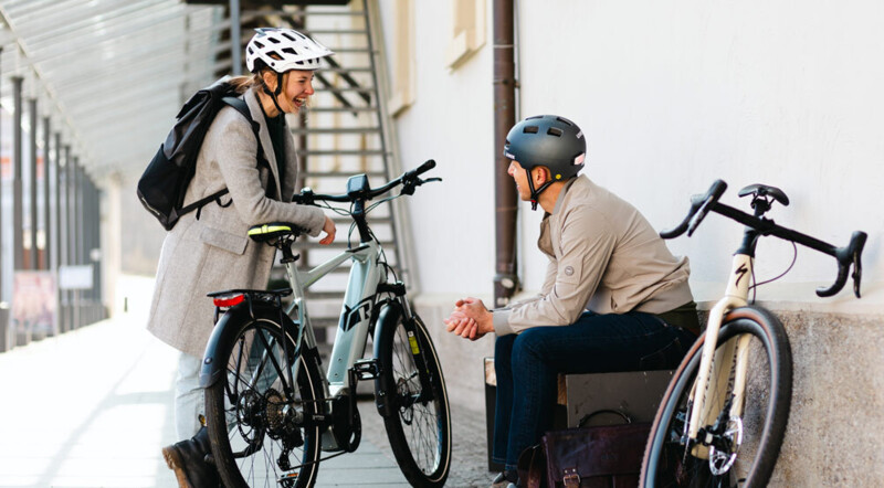 Eine Frau und ein Mann, beide mit Fahrradhelmen, unterhalten sich lächelnd. Die Frau steht neben einem E-Bike, während der Mann auf einer Bank sitzt, neben ihm ein Rennrad.